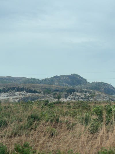 The image depicts a mountain near Kujama, Nigeria. The mountain face is rocky and layered, with sparse vegetation growing on its slopes. Dry, tall grass occupies the foreground. Overhead power lines are visible against the overcast sky.