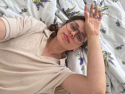 A woman lies on a bed with a white duvet cover printed with purple lavender sprigs. She is wearing round-framed glasses and a beige long-sleeved shirt. Her left arm is raised, with her hand resting on her forehead. The setting is indoors, likely a bedroom. The location is Kőröshegy, Hungary.