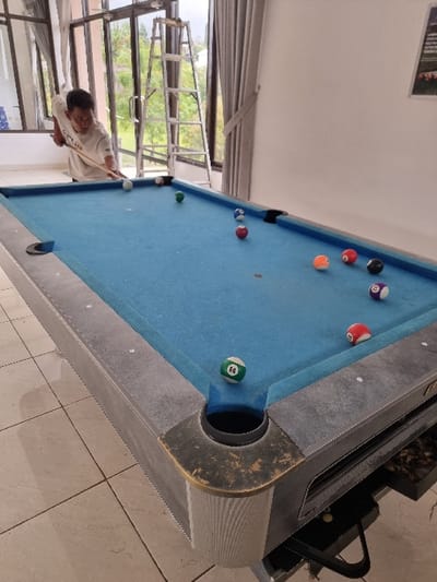 The image depicts an indoor scene in Sindangjaya, Indonesia, featuring a man playing billiards. A light-skinned adult male, wearing a short-sleeved white t-shirt with a dark logo, is positioned on the left side of a blue-felted pool table, bent over and holding a wooden cue stick with both hands. He is in the act of striking a white cue ball.
The pool table, gray with dark corner accents and golden-colored trim on the pockets, occupies the foreground and center of the frame. Multiple colored billiard balls are scattered across the blue felt surface, including a green and white striped ball numbered "14" situated near the lower-right corner pocket. Other balls visible include solid red, orange, black, and blue, as well as striped purple.
In the background, large glass windows with dark frames reveal an outdoor view of green foliage and distant terrain. A silver step ladder stands upright near the central window frame. Light-colored curtains are drawn on either side of the windows. The room features a light-colored tiled floor with a grid pattern. A portion of a framed picture is visible on a white wall in the upper-right background. Natural light illuminates the scene through the windows.