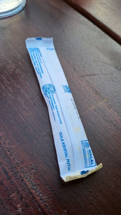 A sugar packet is placed on a wooden surface. The packet is white with blue text and graphics. The text on the packet is in Indonesian and includes information such as "GULA KRISTAL PUTIH" (White Crystal Sugar), "Komposisi: Gula Kristal Putih" (Composition: White Crystal Sugar), and a Halal certification logo. There are also QR codes printed on the packet. The wooden surface appears to be a table with a visible wood grain. The lighting suggests an outdoor or well-lit indoor environment. The image is taken from a slightly elevated angle, focusing on the sugar packet. The specific location of Bireuenmeunasahdayah, Indonesia is not visually discernible from the image itself; this information would be external context.