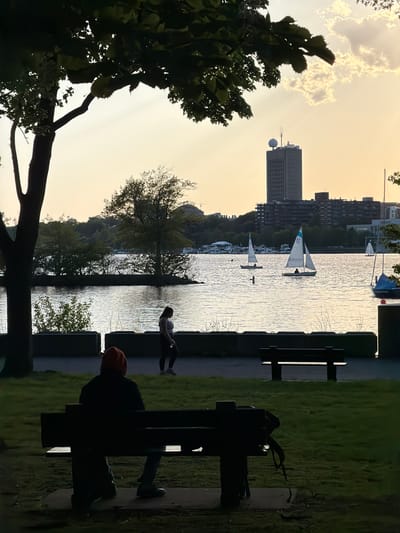 The image shows a view of the Charles River in Boston, United States of America, from a park bench. On the right, a person is sitting on the park bench, and another person is leaning on the edge near the water. Sailboats are on the river, and buildings are visible in the background, including a tall building with a distinctive round structure on top. The sky is bright, indicating sunset or sunrise. Trees frame the left side of the image.