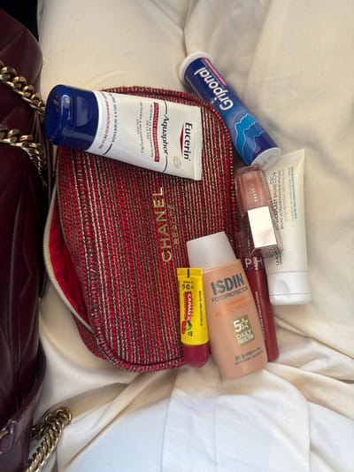 The scene in Aveiro, Portugal, features a collection of personal care and cosmetic products arranged on a light-colored, possibly off-white, textured fabric surface. On the left, a burgundy quilted leather bag with a gold chain strap is partially visible. Centrally, a red textile pouch with prominent gold thread embellishments and "CHANEL BEAUTÉ" embroidered in gold lies partially open, revealing its red interior. Items are scattered across the fabric and near the pouch. These include a white tube of Eucerin Aquaphor Pomada Reparadora, a blue tube of Griponal effervescent tablets, a white tube of La Roche-Posay Cicaplast Baume B5+ ultra-repairing soothing balm, a Fenty Beauty Gloss Bomb Universal Lip Luminizer, a dark red Sephora Collection lip product, a peach-colored bottle of ISDIN Fotoprotector FusionWater Magic Medium SPF 50, and a yellow tube of Carmex Cherry SPF 15 moisturizing lip balm. All objects are stationary.