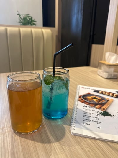 Two beverages in can-shaped glass tumblers are prominently displayed on a light wooden table in what appears to be a restaurant or cafe in Kecamatan Cisaat, Indonesia. The drink in the foreground is an amber-orange liquid, while the one behind it is a vibrant blue with ice cubes and mint leaves, featuring a black bent straw.
To the right of the drinks, a spiral-bound white menu is open, listing "APPETIZER" items. Visible text includes "CHICKEN KARAGE", "CHICKEN SKIN KARACH", "DIMSUM MENTAI" (with Japanese text "通々めんたい" below it), "DIMSUM ORIGINAL", and "GYOZA" (with Japanese text "親子" below it), along with prices such as 27.5, 29, 25, and 30.
The background shows light-colored, padded booth seating and a dark wall or window area. A tissue box is also visible on the table further back. A potted plant with green foliage can be seen behind the seating on the left side of the image. No people are visible in the frame, suggesting a moment before or after dining. The indoor setting is well-lit, indicating daytime.