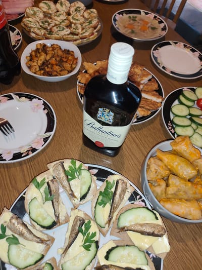 This image shows a table laden with a variety of dishes and a bottle of Ballantine's Finest Scotch Whisky. The spread appears to be part of a meal or gathering.
In the foreground, there are several open-faced sandwiches topped with cheese, cucumber slices, a sprig of parsley, and what appears to be sardines. To the right, a bowl is filled with stuffed cabbage rolls in a rich sauce. Further back, a plate holds pieces of fried food, possibly chicken or fish. Another bowl contains a savory dish with what looks like chicken and vegetables. In the upper part of the image, a plate of appetizer-style bites, possibly bruschetta, is visible, as well as a shallow bowl of clear soup with shredded meat and carrots. A bottle of Coca-Cola is partially visible on the left. The setting is a wooden table.
The lighting suggests it could be daytime, indoors. There are no people visible in the image. The overall impression is of a home-cooked meal with a generous assortment of food. Visible text on the bottle includes "Ballantine's Finest Blended Scotch Whisky" and "Scotland".