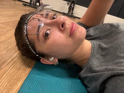 An individual is depicted lying on their back on a teal exercise mat in what appears to be an indoor fitness facility. The subject, a female with dark hair, is wearing a grey short-sleeved t-shirt and a black Adidas headband with white text pushed up onto her forehead. Clear-framed glasses with round lenses are resting on the headband. Her face exhibits a neutral expression with a subtle sheen on the skin. Multiple piercings are visible on her left ear, including a white earbud-like object and a small hoop earring with a light-colored bead. The floor is light-colored wood, and in the background, partially obscured and out of focus, are dark-colored stationary exercise bicycles. The specific city of Komchén, Mexico, is not discernible from the visual information provided within the image.