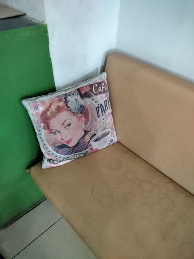 A vintage-style throw pillow rests on a light brown upholstered bench, which is positioned in the corner of a room. The room features a bright green wall on the left, meeting a lighter, possibly white or off-white, wall on the right. The floor in the lower left of the frame appears to be tiled in a light, possibly gray or beige, color with a visible grout line.
The pillow is rectangular with a white border. Its design features a prominent illustration of a woman with red hair styled in an updo, looking to the side with a knowing or playful expression. Her lips are red, and she wears a patterned dress or top. Next to her, a steaming coffee cup is depicted, below which French text reads "Une Saveur Chic et..." (A Chic Flavor and...). More French text is visible at the top: "Le Cafe" and "PARIS," partially obscuring an image of the Eiffel Tower. Along the bottom curve of the pillow's design, "POUR LA PARISIENNE" (FOR THE PARISIAN WOMAN) can be seen.
The bench itself is a solid, simple design, showing some subtle texture and a few faint marks or discolorations on its surface, suggesting wear. The green wall is uniformly painted, while the lighter wall shows minor imperfections. The scene is static, depicting an unoccupied corner, possibly in a cafe, waiting area, or home in Dramaga, Indonesia, given the provided context. The overall impression is one of a quiet, decorative indoor space.