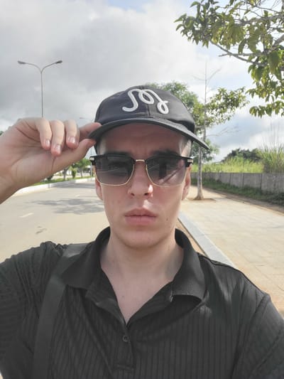 A man wearing a black baseball cap with a white logo, black sunglasses with gold trim, and a black collared shirt stands on a paved walkway. He is holding the brim of his cap with his left hand. The man is looking directly at the camera. The background shows a road, trees, a concrete wall, and a cloudy sky. The location is identified as Bo Y, Vietnam.