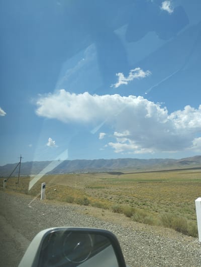 The image captures a view from inside a vehicle looking out towards a vast, arid landscape under a bright blue sky with scattered clouds. In the foreground, a car's side mirror reflects a portion of the interior and the road ahead. The road itself appears to be gravel, with the shoulder marked by white posts. Beyond the road, the terrain transitions into dry, yellow-green vegetation, characteristic of a steppe or semi-desert environment. In the middle ground, a line of trees and buildings suggests a settlement or village, stretching towards a range of low, rolling mountains in the background. A solitary utility pole with wires is visible on the left side of the frame, indicating infrastructure in the area. The scene is consistent with the rural outskirts of Forish QFY, Uzbekistan, known for its expansive natural landscapes and agricultural settlements.