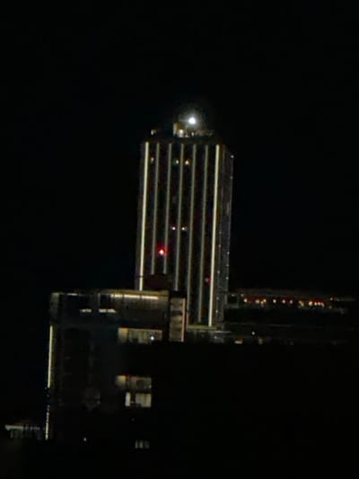 The image displays the Mercury Towers, a prominent skyscraper located in St. Julian's, Malta, viewed at night. The scene is consistent with a perspective from the nearby city of San Ġwann.
The central subject is the tall, slender skyscraper, starkly silhouetted against a completely dark night sky. Its facade is prominently illuminated by numerous vertical white light strips extending along its full height. A distinct, bright spherical light source, consistent with either the moon or a powerful rooftop luminaire, is positioned directly on the building's apex. Multiple small red aviation obstruction lights are visible on various upper levels of the tower.
Below and to the left of the main tower, a broader, lower building complex, either integrated or immediately adjacent, exhibits scattered illuminated windows, indicating interior lights. In the distant background, a blurred horizontal line of numerous small, yellow-orange lights suggests a further urban landscape. No individuals or specific actions are discernible within the frame. The overall environment is one of an urban nighttime setting.