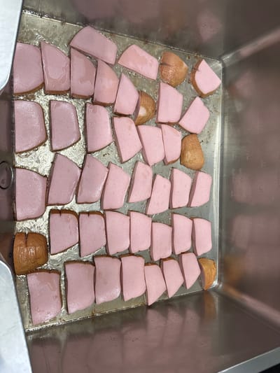 A top-down view shows a stainless steel container filled with slices of meat. The meat slices are primarily pink with a brown edge. Some of the slices are fully brown. The container is partially filled with oil. The container has volume markings on the side. There is no visible indication of the location in Neumünster, Germany. There are no people visible.