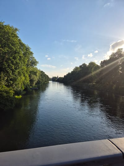 This image depicts a wide river flowing through a city, flanked by lush green trees on both sides. The sky is a bright blue with a few scattered clouds, and the sun is casting visible rays through the trees on the right side of the image. In the distance, a few buildings, including what appears to be a church tower, are visible. The water in the river is dark and reflects the trees and sky, with ripples indicating a gentle current. The foreground shows the edge of a dark-colored railing, suggesting the photo was taken from a bridge.
This view is characteristic of the Isar River in Landshut, Germany. The specific perspective, with the river flowing from the viewer's perspective towards a distant cityscape, is typical of vantage points along the riverbanks or bridges within the city. The presence of dense trees lining the river is also a common feature of the Isar's path through Landshut. The distant church tower is likely part of the historic Altstadt (Old Town) of Landshut.