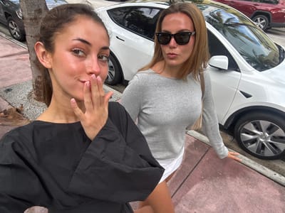 Two adult females are positioned on a reddish-pink concrete sidewalk in Miami Beach, United States, under natural daylight. The female in the foreground on the left, with light brown hair pulled back, wears a black long-sleeved top. Her right hand, featuring light pink manicured fingernails, is raised with fingers touching her lips as she looks directly at the camera. The female in the background on the right, with light brown hair down, wears dark sunglasses, a light grey long-sleeved top, and white shorts. She carries a light-colored shoulder bag and has slightly pursed lips, directing her gaze towards the camera. Behind the subjects, a white Tesla Model Y is parked parallel to the street curb, partially obscuring a dark red SUV. A tree trunk is visible on the left between the women and the parked vehicles.