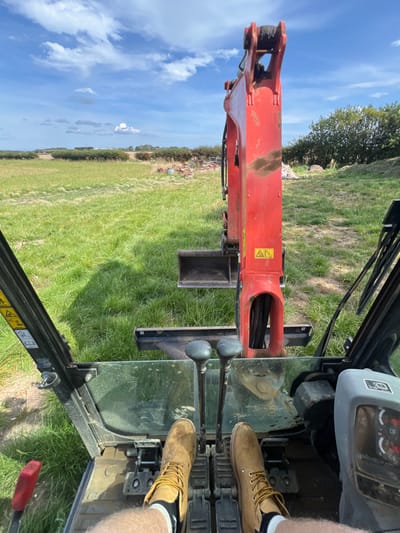 An operator sits in the cab of a mini-excavator with their feet on the foot pedals. The operator is wearing tan boots with black socks. The excavator's boom extends outward, and a small bucket attachment is visible. The background consists of a grassy field and a blue sky with scattered clouds. The photo is taken from an elevated perspective within the cab.