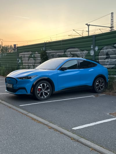 A bright blue Ford Mustang Mach-E electric vehicle is parked in a lot with asphalt paving and white parking lines. The car is positioned facing left, slightly angled towards the viewer. It has black trim around the wheel wells and along the lower body. The license plate reads "HWL 87E". In the background, a green sound barrier with graffiti is visible, behind which are train tracks and overhead power lines. The sky is a pale orange and yellow, suggesting either sunrise or sunset. There are no people or animals present in the image. The environment appears to be an urban or semi-urban setting, likely adjacent to a railway line, within Wustermark, Germany.
