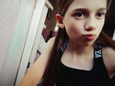 The image shows a close-up selfie of a young girl with long brown hair, looking directly at the camera with her lips pursed. She is wearing a black tank top with a white logo on the strap. The background is blurry but appears to be indoors, possibly a room with a clock on the wall and some clothing. The exterior wall of a building with a textured surface is visible on the left side of the frame. This image was taken in Radzymin, Poland.