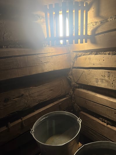 The image depicts an interior view of an attic or a similar unfinished space, characterized by its exposed wooden beams and planks. The rough-hewn wood, some with visible knots and grain, suggests an aged or rustic construction. The scene is dimly lit, with a bright light source originating from the left, possibly a window or a lamp, which casts distinct shadows and illuminates the wooden surfaces.
On the left side of the image, a wooden slatted structure is visible, partially obscured by a hazy, luminous glow. This could be a vent or a rudimentary window covering. In the right portion of the frame, two metal buckets are prominently featured, one of which is partially filled with a clear liquid, reflecting the ambient light. The buckets appear to be hanging or placed on the wooden beams.
The texture of the insulation or padding between the wooden beams is also noticeable, appearing fibrous and somewhat disheveled. The overall impression is one of a utilitarian or storage space, perhaps undergoing repair or simply left in its raw state. There is no visible text in the image, nor are there any people present. The lighting suggests it could be daytime, with light filtering in, but the enclosed nature of the attic makes it difficult to determine the exact time of day or weather conditions outside. The location context of Аргамак, Russia, implies a cold climate, which might explain the presence of insulation, though this is speculative based solely on the image. The scene evokes a sense of quiet utility, with the buckets possibly being used to collect water, hinting at a potential leak or a need for temporary water storage.