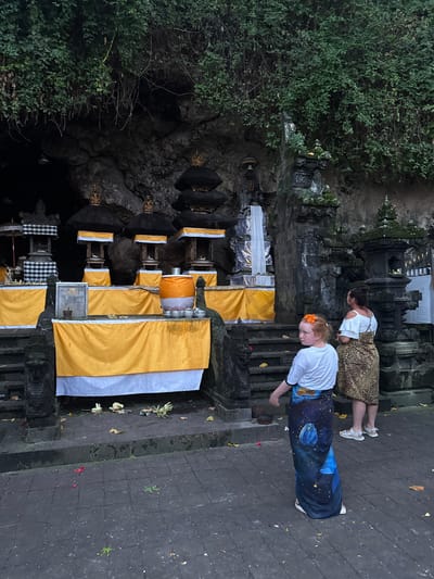 In Dawan, Indonesia, at the Goa Gajah (Elephant Cave) archaeological site, a woman and a young girl stand on a stone pathway in front of a series of offerings. The girl, facing away from the camera, wears a white t-shirt and a long, dark blue sarong patterned with blue flowers and stars. She has an orange flower in her hair. The woman, to her right and slightly behind, wears a white off-the-shoulder top and a leopard-print sarong.
The offerings are displayed on tiered stone platforms draped with yellow and white cloth. Several elaborately decorated shrines with black, tiered roofs topped with gold ornaments are visible. A large orange and white cylindrical object, possibly a donation box or offering, sits on one of the platforms, surrounded by small silver cups. In the background, the entrance to a cave is visible, partially obscured by lush green foliage cascading down the rocky hillside. The ground is paved with dark square tiles, and scattered fallen leaves and flower petals indicate a natural outdoor setting. Stone carvings of mythical creatures or guardians are present on either side of the pathways and stairs. The overall scene suggests a spiritual or cultural site.