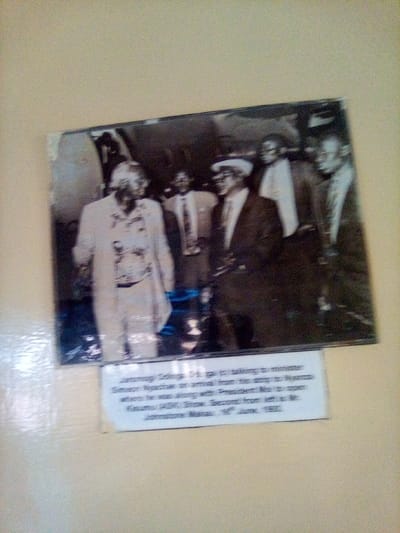 A black and white historical photograph, approximately 20cm x 15cm, is mounted on a plain, light-tan wall in the South Sakwa ward, Kenya. The photograph's upper-left corner shows minor physical damage. The image depicts five or more adult males in an outdoor setting, with a large, dark, indistinct structure, possibly an aircraft fuselage, visible in the background. The prominent figure on the far left, identified by an accompanying caption as Jaramogi Oginga Odinga, wears a light-colored, patterned long-sleeved shirt and light trousers, and is positioned facing slightly right. To his right, Minister Simeon Nyachae, dressed in a dark suit, light shirt, tie, and a light-colored fedora, smiles while looking left towards Odinga, seemingly engaged in conversation. Behind Nyachae, Mr. Johnstone Ndiritu, also in a dark suit, light shirt, and tie, is visible and smiling. Other male figures are present in the blurred background. A white label with black text adhered directly below the photograph provides detailed context: "Jaramogi Oginga Odinga (1st gal) talking to minister Simeon Nyachae on arrival from his airstrip to Nyanza where he was along with President Moi to open Kasumu (ASK) Show. Second from left is Mr. Johnstone Ndiritu. 18th June, 1991." This text specifies the event as Odinga conversing with Nyachae upon Nyachae's arrival from an airstrip in Nyanza on June 18, 1991, during an occasion when President Moi was also present to open the Kisumu (ASK) Show.