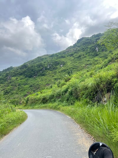 An asphalt road curves to the left, bordered by tall green grass and vegetation on the right. To the left of the road, the terrain rises steeply, covered in lush green trees, shrubs, and patches of exposed rock. In the distance, a mist-shrouded mountain looms under a cloudy sky. The reflection of a person wearing a black shirt and a necklace is visible in a dark, circular mirror in the bottom right corner of the image. The scene is located on a road in Lùng Tám Commune, Vietnam.