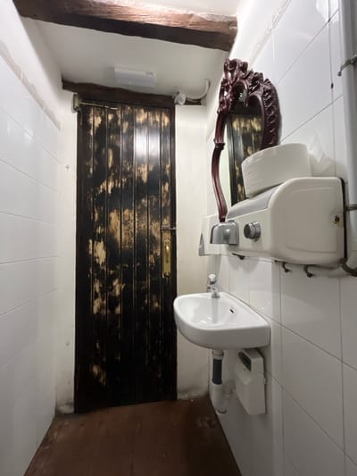 The image depicts a small bathroom in a building located in Agüimes, Spain (latitude: 27.907, longitude: -15.447). The bathroom has white tiled walls and a brown tiled floor.  On the right side of the room is a white sink with a chrome faucet. Above the sink is a white hand dryer mounted to the wall. Next to the hand dryer is a decorative, ornate mirror, partially obstructed by a white, rectangular box containing paper towels. On the left side of the room is a wooden door with a brass handle, leading to another room. The door is dark brown, with a weathered, distressed finish. There is a small, wooden beam running across the ceiling of the room.