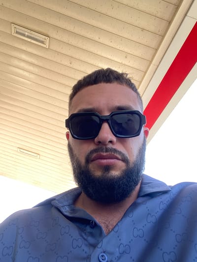 A man wearing black sunglasses and a blue patterned button-up shirt is positioned in the center of the frame, looking upwards towards the camera. The background consists of a textured ceiling and a white and red stripe. A rectangular light fixture is visible on the ceiling. The image appears to have been taken outdoors in a location with overhead cover, possibly at a gas station or a covered walkway. The man has a beard and short hair. The overall scene is a close-up portrait against an architectural backdrop.