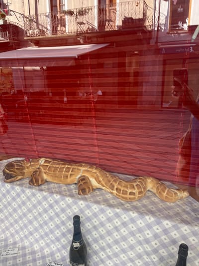 The image shows a window display of a bakery or food shop. In the foreground, a bread sculpture shaped like a crocodile or lizard lies on a checkered tablecloth. The bread is golden brown and has a textured surface resembling scales. Behind the bread sculpture, a red pleated blind is visible, and further back, through the glass, there are reflections of a street with balconies and buildings. A bottle of sparkling wine stands on the tablecloth to the right of the bread sculpture, and there are two small signs on the tablecloth to the left and bottom. The scene is likely located in the city of Aosta, Italy, as indicated by the Italian text on the signs.
