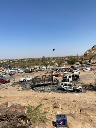 The image shows a wide shot of a large, open area in Tayba Alhasanab, Sudan, characterized by a terrain of sandy soil and scattered rocks. The area is littered with the remains of burnt-out vehicles, including cars, trucks, and SUVs. Some of the vehicles are partially charred, while others are completely destroyed. The remnants of the vehicles are scattered haphazardly across the open space, with tire tracks and debris creating a chaotic scene. A single black bird is soaring in a clear blue sky above the scene. In the distance, a small town can be seen with low-rise buildings surrounded by sparse vegetation. A lone Pepsi crate sits on a large rock in the foreground. The image suggests a aftermath of a conflict or destruction in the area.