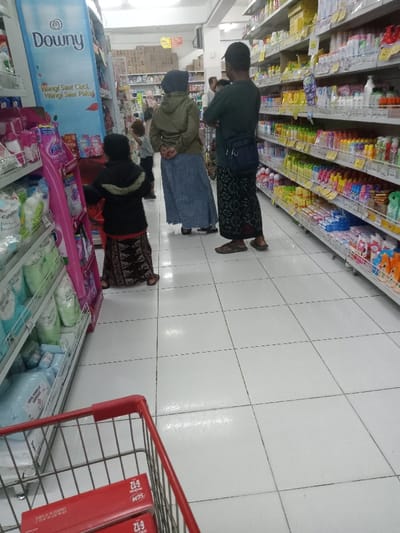 The image depicts an interior view of a supermarket aisle in Nganjuk, Indonesia. The floor is comprised of white ceramic tiles with dark grout lines, and the space is illuminated by overhead artificial lighting.
In the mid-ground, three individuals are prominently visible from behind: two adults and one child. A female adult on the left wears a grey hijab, a khaki-colored long-sleeved top, and a long blue denim skirt, with her hands clasped behind her back. To her right, a male adult wears a dark cap, a dark green short-sleeved shirt, a dark patterned sarong, and dark flip-flops, with his arms crossed and a dark sling bag draped over his left shoulder. A young child stands in front of the female adult, wearing a dark jacket and a dark patterned sarong, appearing to reach towards a pink display stand on the left aisle. A fourth adult is partially visible further in the background between the two main adults, wearing a white shirt.
The aisle is bordered by metal shelving units on both sides, stocked with various merchandise. The left side features a prominent blue vertical advertisement for "Downy" fabric softener with Indonesian text. Below this, shelves display household products in light green and blue packaging, alongside a pink promotional display. The right side of the aisle is densely packed with numerous colorful bottles and packages, primarily personal care items and toiletries.
In the immediate foreground, the red handle and front metal basket of a shopping cart are visible. The cart contains two red boxes, each labeled "SCM 6-12".