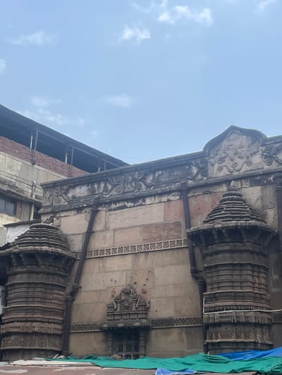 This is a vertical, eye-level, outdoor shot of an ancient stone building in Ahmedabad, India. The building features intricate carvings and architectural details, including ornate pillars and decorative friezes. The stone appears weathered and has a reddish-brown hue in some sections, contrasting with areas of grey stone. A dark, possibly rusted, pipe runs vertically down the facade. In the foreground, a bright green tarp is draped over some unseen objects, suggesting ongoing work or protection. The sky above is a pale blue with scattered white clouds. The architectural style points to a historical structure, likely a temple or mosque, given the region.