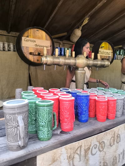 In Óbidos, Portugal, a stall at a medieval fair is presented. A woman with dark hair, wearing a light-colored dress, stands behind a bar with taps for drinks. In front of her, a variety of ceramic mugs in different colors, including red, green, blue, and gray, are displayed. Behind the bar, two wooden barrels are visible, one labeled "Cerveja Artesanal" and the other "Hidromel." The stall is set up outdoors, with a wooden roof and canvas backdrop. The overall scene depicts a vendor selling artisanal beverages at a historical reenactment or fair.
