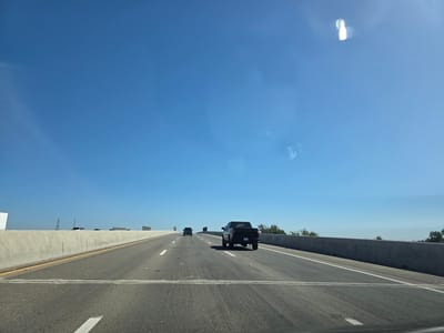 A clear, blue sky stretches above a highway in West Sacramento, United States of America. A black pickup truck is driving in the right lane, and a smaller truck is in the left lane. The highway has concrete barriers on both sides. In the distance, a few trees and power lines are visible. The sun is shining, casting shadows on the road. There are some lens flares in the sky.