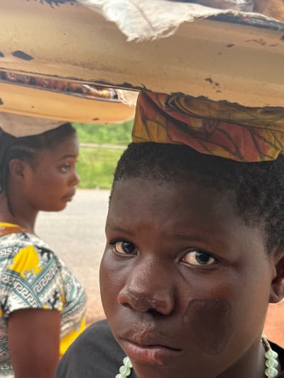 In Umuoye, Nigeria, two young females are featured. The foreground is dominated by a close-up of a young female's face, looking directly at the camera with a somber expression. She has dark skin and her head is adorned with a patterned cloth. A smudge of dark pigment is visible on her right cheek. She is wearing a dark t-shirt and a necklace of light green beads. Behind her, slightly out of focus, is another young female in profile, also carrying an object on her head. She has braided hair and is wearing a vibrant, patterned shirt with yellow and blue elements. The background suggests an outdoor setting with a blurred green area and a paved surface. The two individuals appear to be walking or standing in close proximity, possibly as part of a market scene or daily activity in the community.
