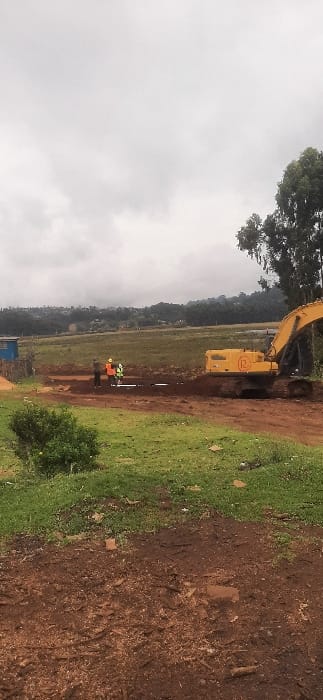 A construction scene under a cloudy sky shows a yellow excavator working alongside a dirt road. Two figures in orange and green vests stand nearby, possibly workers overseeing the operation. The landscape includes a grassy field stretching out behind them, with some trees visible in the background. The foreground consists of a mix of grass and exposed dirt, suggesting recent excavation or ground disturbance.