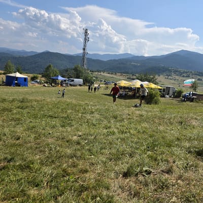 The image depicts an outdoor gathering on a grassy hillside, likely in Ravnogor, Bulgaria, given the mountainous terrain and the presence of a Bulgarian flag. In the foreground, the grass is dry and appears to have been recently cut in places. Scattered throughout the scene are several tents and canopies, set up for an event. One large yellow canopy, branded with "AQUA" and "Sok" logos, provides shade for a group of people seated at tables. Another smaller blue tent is visible to the left, with equipment, including what appears to be a speaker, set up inside.

Several individuals are present in the image. Two young boys are seen in the middle ground, one sitting and the other standing. Further back, several people are gathered under the yellow canopy, some seated and others standing. A young person in a red shirt is running through the grass in the middle right of the frame, with a soccer ball nearby. To the far right, a Bulgarian flag, consisting of white, green, and red horizontal stripes, is planted near a table with some items on it. A white van and a small caravan are parked behind the yellow canopy.

In the background, a tall telecommunications tower stands out against the sky, which is partly cloudy with dramatic, billowy white clouds. The landscape behind the gathering is characterized by rolling, forested hills and mountains, typical of the Rhodope Mountains region where Ravnogor is located. The overall scene suggests a casual outdoor event or festival.