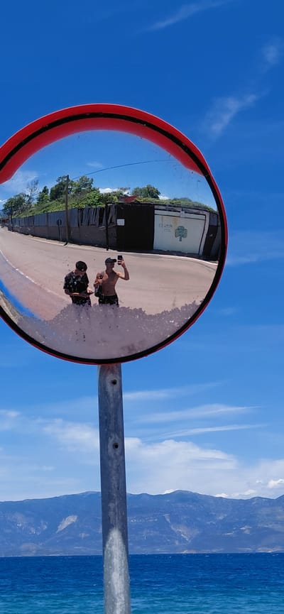 A vertical, slightly angled shot captures a convex traffic mirror reflecting a street scene. The mirror, with a red rim and a grey pole, is positioned against a bright blue sky with scattered clouds. The reflection shows two young men, one holding a smartphone to take a picture, standing on a paved road. Behind them is a fence and buildings. The lower part of the mirror has a weathered appearance with white debris. In the background, a vast blue sea meets a range of distant, muted mountains under the expansive sky. The scene is set in the Municipal Unit of Aegio, Greece.
