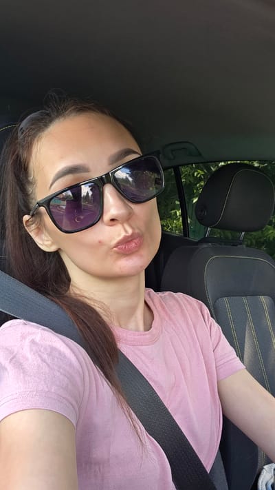 A young woman with dark hair pulled back in a ponytail is sitting in the driver's seat of a car, wearing sunglasses and a pink t-shirt. She is looking at the camera with her lips pursed. A seatbelt is visible across her chest. The car's interior, including a headrest, is visible in the background. The scene suggests she is in a car, potentially driving on a road, given the reflection in her sunglasses which appears to show trees and a road. The location is specified as Старошареево, Russia.