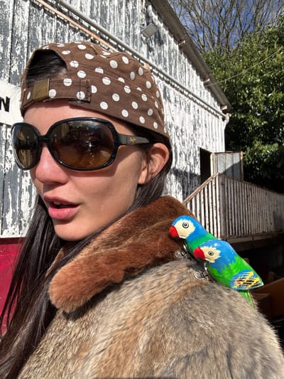 A close-up outdoor portrait in Neuilly-Plaisance, France, features a female subject positioned slightly right of center, looking towards the left with her mouth slightly open. She wears a brown fabric cap with white polka dots, worn backward, and large, dark, oval-shaped sunglasses with reflective brown-tinted lenses (Maui Jim branded). Her long dark hair is visible on her left side. She is dressed in a fur or faux fur coat, distinguished by a darker brown collar and a lighter, mottled brown body. Two small, brightly painted wooden parrot brooches, featuring blue heads, white faces with red beaks, and green bodies, are pinned to her right shoulder.
The background consists of a weathered wooden building with vertical planks, predominantly white with significant peeling paint revealing darker wood. A white outdoor light fixture is mounted on the building's wall. To the right, a wooden staircase and railing lead to an open doorway on an elevated section of the structure. Behind the building on the right, green tree foliage is visible against a clear blue sky, indicating strong natural sunlight.