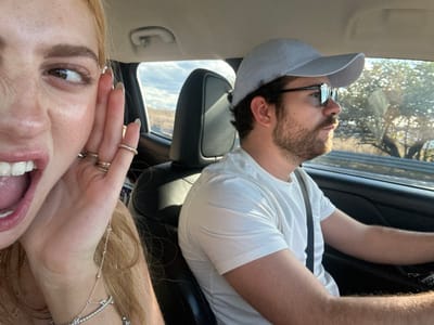 Upside-down selfie featuring a man and a woman inside a car. The man wears a white shirt, baseball cap, and glasses. He is seated in the driver's seat. The woman, with blonde hair and several rings, is in the passenger seat, displaying an open mouth with visible teeth and tongue. A window shows an outdoor scene. The car interior includes a black headrest and a light-colored dashboard.
