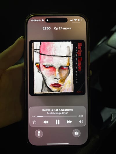The image shows a close-up of a person's left hand holding a smartphone, displaying a music player interface. The phone has a gold-colored frame and a notch at the top. The screen shows the current time as 22:00 and the date as "24 июня" (June 24th) in Cyrillic script. The music player displays album art featuring a stylized portrait of a person with dramatic makeup, and the song title "Death Is Not A Costume" by "MetaManipulator". Below the album art are playback controls, including a progress bar indicating 0:40 played and a total duration of -4:13. There is also a star icon for favoriting, skip forward and backward buttons, a volume icon, and a flashlight icon. The lower right corner of the screen shows a camera icon. The background is dark and out of focus, suggesting the photo was taken in low light conditions, possibly indoors or at night. The phone is positioned on what appears to be a car's dashboard or seat. There is no visible indication of the city of Sękocin Stary, Poland.
