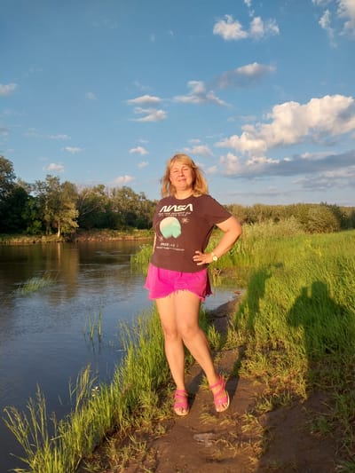 A woman with blonde hair and fair skin, wearing a dark t-shirt with a NASA logo and pink shorts, stands on a grassy bank next to a body of water. She is facing the camera with her right hand on her hip and wearing pink sandals. The water appears to be a river or a large pond, with trees and vegetation on the opposite bank and a grassy field to her right. The sky is blue with scattered white clouds, and the sun is setting, casting long shadows. The scene suggests a natural outdoor environment, possibly in a park or a rural area near the city of Chkalov, Russia, given the context.