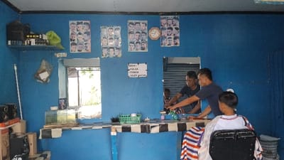 This image captures the interior of a modest barbershop, likely in Panjalu, Indonesia. The walls are painted a vibrant blue, and the space is simply furnished.
In the foreground, a young boy with short hair sits in a barber's chair, draped with a red, white, and blue striped cape. He is facing away from the camera. Behind him, a male barber, wearing a dark blue t-shirt, is actively cutting his hair. The barber is focused on his task, with his hands positioned around the boy's head. Another man, also wearing a dark t-shirt, is visible in the large mirror directly in front of the barber, possibly waiting or assisting. The mirror reflects a glimpse of the outdoor environment.
The barbershop is decorated with several posters displaying various men's hairstyles, suggesting options for customers. A round wall clock is also visible on the blue wall between two of these posters. On the left side of the room, an open window lets in bright daylight, revealing green foliage outside. Below this window, a small aquarium sits on a checkered shelf, which also extends across the main counter where the haircut is taking place. Various barber tools and spray bottles are arranged on this counter. On a shelf high up in the left corner, there are speakers, a green bag, and other items.
A white sign, handwritten with black marker, is prominently displayed on the blue wall, stating the services offered:
* "SEDIA:" (Available:)
* "PULSA" (Mobile credit/top-up)
* "TOKEN" (Electricity token)
* "SILET ANGKAT" (Razor blades/shaving supplies)
The bright lighting suggests it is daytime. The overall ambiance indicates a local, unpretentious setting, characteristic of a community barbershop.