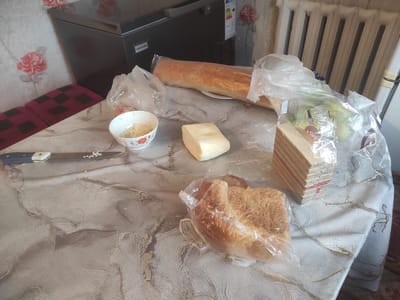 A table covered with a patterned tablecloth is set with food items. A long baguette is wrapped in plastic, next to a bowl of shredded cheese. A block of cheese sits in the center of the table, and a package of wafers is to the right. A knife with a white handle and a partially chopped baguette in a plastic bag are also visible on the table. In the background, a refrigerator with a label that reads "AVANGARD" is present, along with a radiator and a floral-patterned wallpaper. The scene appears to be indoors, likely in a residential kitchen in Nekrasovka, Kazakhstan.