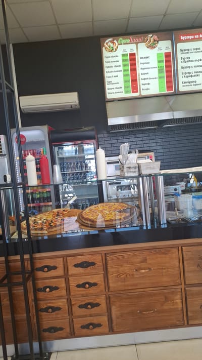 The image shows the interior of a fast-food establishment in Plovdiv, Bulgaria. A menu board displays food items such as gyros, souvlaki, burgers, and more, with prices listed in Bulgarian currency (Leva). A refrigerator case houses Coca-Cola products and other beverages. A display case showcases pizzas. A wooden cabinet with drawers is positioned to the right. The scene appears to be a typical fast-food setting.