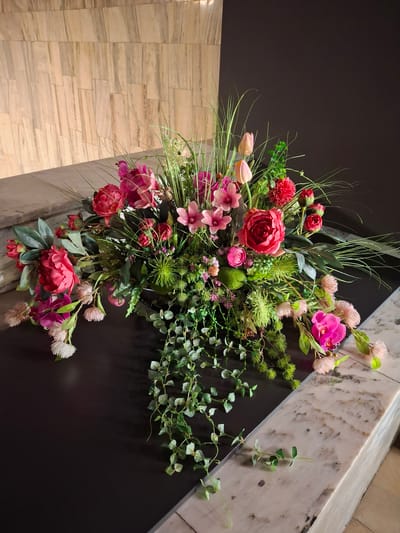 The image shows a floral arrangement on a dark surface, possibly a countertop or table, with a marble edge. Behind the arrangement is a wall with a vertical patterned texture, likely stone or tile. The floral arrangement is lush and features a variety of flowers in shades of pink, red, and fuchsia, along with greenery and wispy grass-like elements. Several pink tulips are visible, as are large peony-like flowers, smaller pink blossoms, and a bright pink orchid. A cascading vine with small green leaves trails down the side of the arrangement. The scene is well-lit, casting soft shadows. The location is described as being in the city of Pila, Poland, within a building.