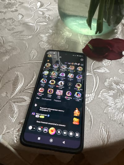 A smartphone with a dark interface is displayed on a tablecloth with a floral pattern. The screen shows various profile pictures of users, likely from a social media or chat application, with names and follower counts. Some of these names are in Cyrillic script, indicating a Russian-speaking user base. At the top of the screen, the time "16:52" is visible. The phone is oriented in portrait mode. In the background, a glass vase with green stems and a red rose petal are partially visible. The overall scene suggests a casual indoor setting. There is no specific indication of the location being Tumanyan, Armenia.