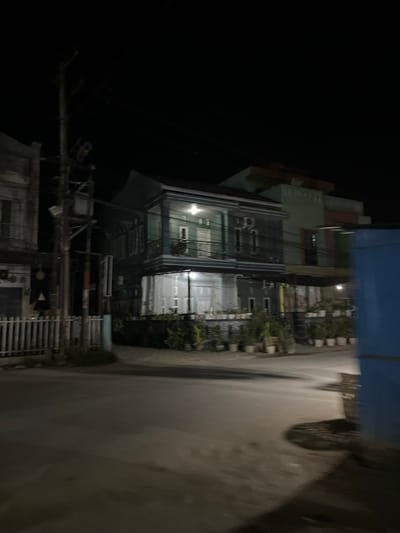 A nighttime street view in Landuh, Indonesia, shows a two-story building with green and gray siding. The building has a balcony on the second floor and is illuminated by interior lights. Several potted plants are arranged along the front of the building. To the left, a white fence borders a sidewalk and a utility pole with wires. A blue structure is visible on the right edge of the frame. The street in the foreground is paved and appears wet, reflecting ambient light. The overall scene depicts a quiet urban residential or commercial area at night.