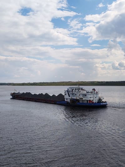 A barge carrying a load of dark material, likely coal, is traveling on a wide river. The barge is composed of two sections: a long, flat-bottomed portion filled with the cargo and a smaller, multi-story tugboat attached to the rear. The tugboat is white and blue and has the name "OKCKMM-57" written on its side. The river is relatively calm with small ripples on the surface. A line of trees can be seen on the distant shore. The sky is partly cloudy with patches of blue visible between the clouds. While the picture does not mention the name of the city where this picture was taken, the barge’s registration number states that it is from Russia. There is no Pruzhinino in Russia.