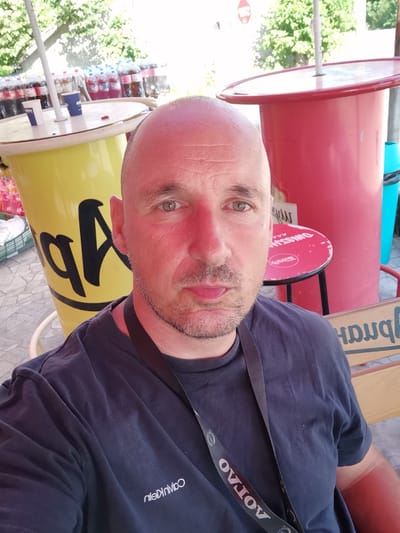 A man with a shaved head and a light beard takes a selfie in front of a yellow cooler and a red stand. He is wearing a dark blue t-shirt with the Calvin Klein logo visible on the left side of his chest. A black lanyard with white lettering is around his neck. In the background, there are rows of bottled drinks, trees, and a red cooler. A small red table with a black base is to the right of the man. A blue trash can is visible behind the red cooler. The flooring appears to be paved with hexagonal tiles. The scene is outdoors, likely in a public area. The specific location in the city of Dralfa, Bulgaria cannot be determined from the visual information alone.