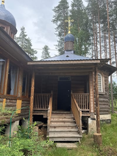 The image shows a wooden church in Nenyamyaki, Russia. The church has a dark blue dome topped with a golden cross and is constructed from logs. A wooden porch with stairs leads to a dark door. To the left of the entrance, there are several windows with white curtains. The church is surrounded by tall pine trees and green vegetation. The sky is overcast.