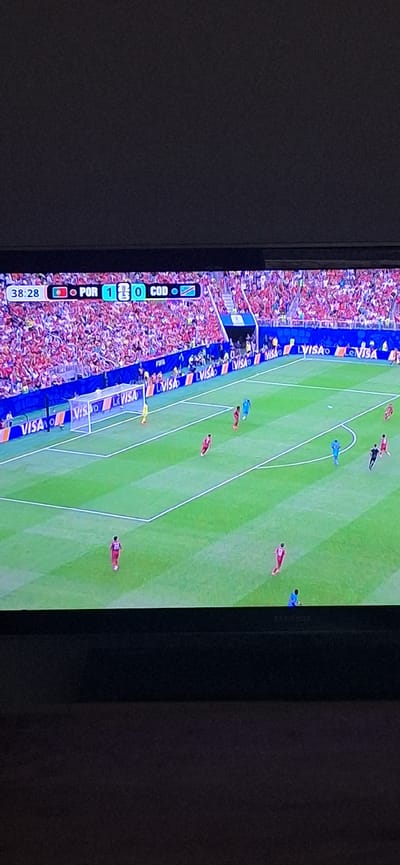 The image shows a screen displaying a soccer match. A score graphic indicates the time is 38:28 and the score is Portugal (POR) 1 - 0 Congo (COD). The game is taking place in a stadium with a large crowd in the background. The field is green, with white markings. There are several players on the field wearing red and blue uniforms. Advertising boards with the VISA logo are visible around the field. The image is taken from an angle, likely from another screen or device. No details are provided about the city of Studena, Bulgaria.